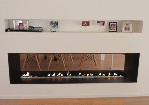 A sleek, modern ethanol bio-fuel fireplace built into a clean white wall, featuring a linear burner with bright yellow-orange flames behind a frameless glass screen. The room has minimalist decor, a light-gray sofa, and soft natural lighting for a contemporary ambiance.