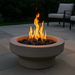 A modern round fire pit constructed from smooth gray concrete, with vibrant orange flames rising from black lava rocks. It’s placed in an outdoor patio area with wooden decking, surrounded by minimalist outdoor seating and ambient evening lighting.