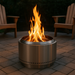 A cylindrical stainless steel smokeless fire pit on a paver patio, with bright orange flames rising from wooden logs. Two wooden Adirondack chairs face the fire, surrounded by greenery in a cozy backyard setting at dusk.