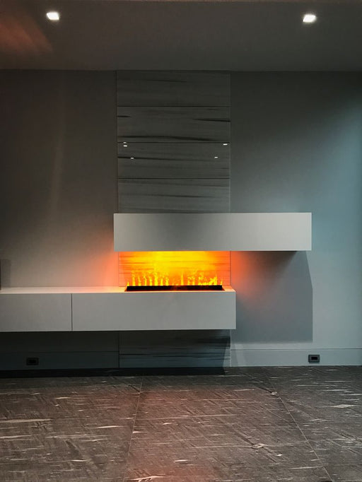 A recessed linear water-vapor fireplace with a matte black frame built into a light gray wall. Ultrasonic mist rises from a narrow burner slot, illuminated by warm orange LED lights to mimic realistic flames above a bed of crystal embers, in a minimalist, modern living space.
