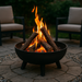 A round wood-burning fire pit made of rusted steel with stacked logs ablaze, placed on a stone patio surrounded by Adirondack chairs. The scene captures a cozy backyard gathering at dusk with warm ambient lighting and trees in the background.