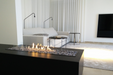 A sleek, modern ethanol bio-fuel fireplace built into a clean white wall, featuring a linear burner with bright yellow-orange flames behind a frameless glass screen. The room has minimalist decor, a light-gray sofa, and soft natural lighting for a contemporary ambiance.