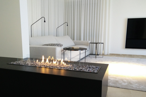 A sleek, modern ethanol bio-fuel fireplace built into a clean white wall, featuring a linear burner with bright yellow-orange flames behind a frameless glass screen. The room has minimalist decor, a light-gray sofa, and soft natural lighting for a contemporary ambiance.